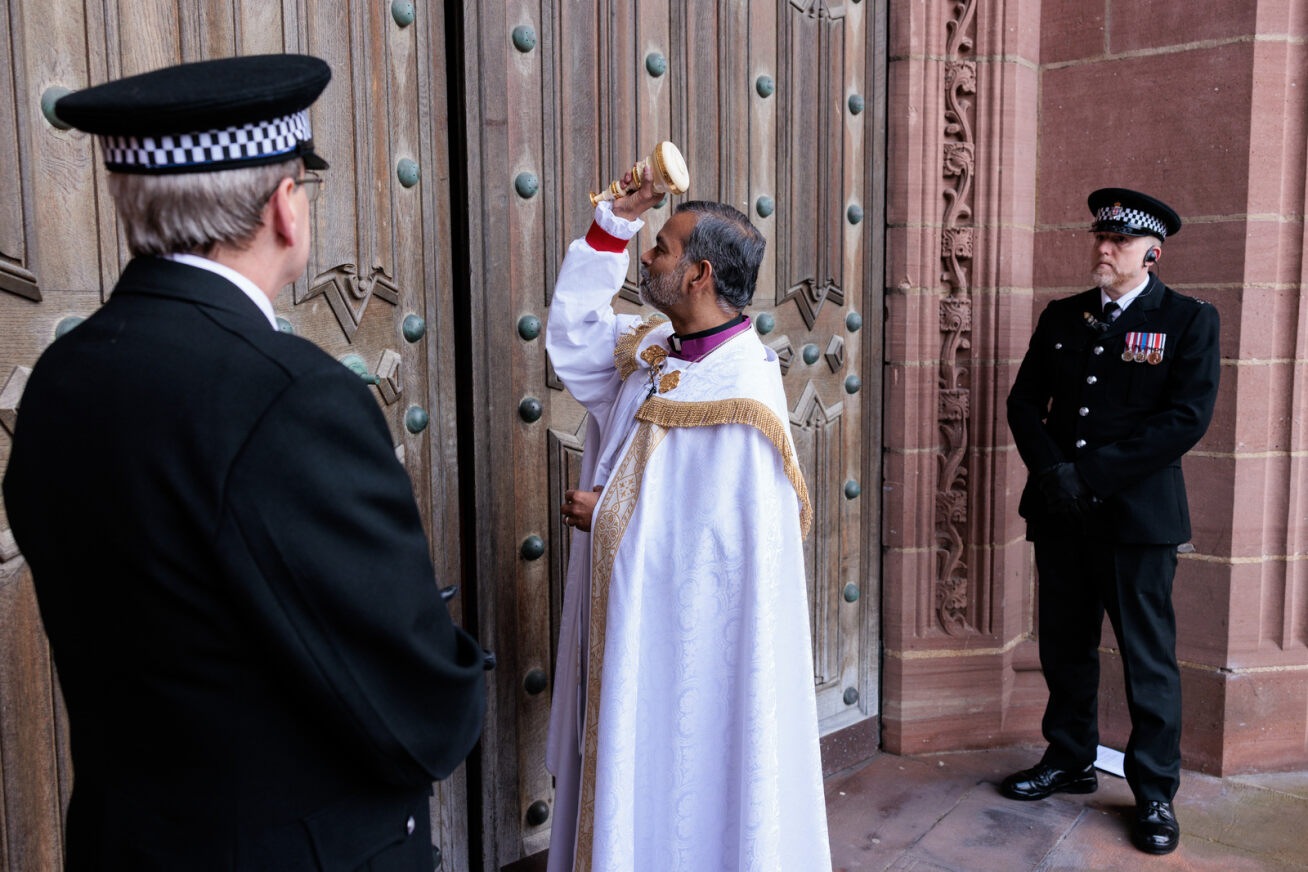 Bishop John in ceremonial robe uses a commemorative mallet to knock on thew great west doors of Liverpool Cathedral while two constables look on