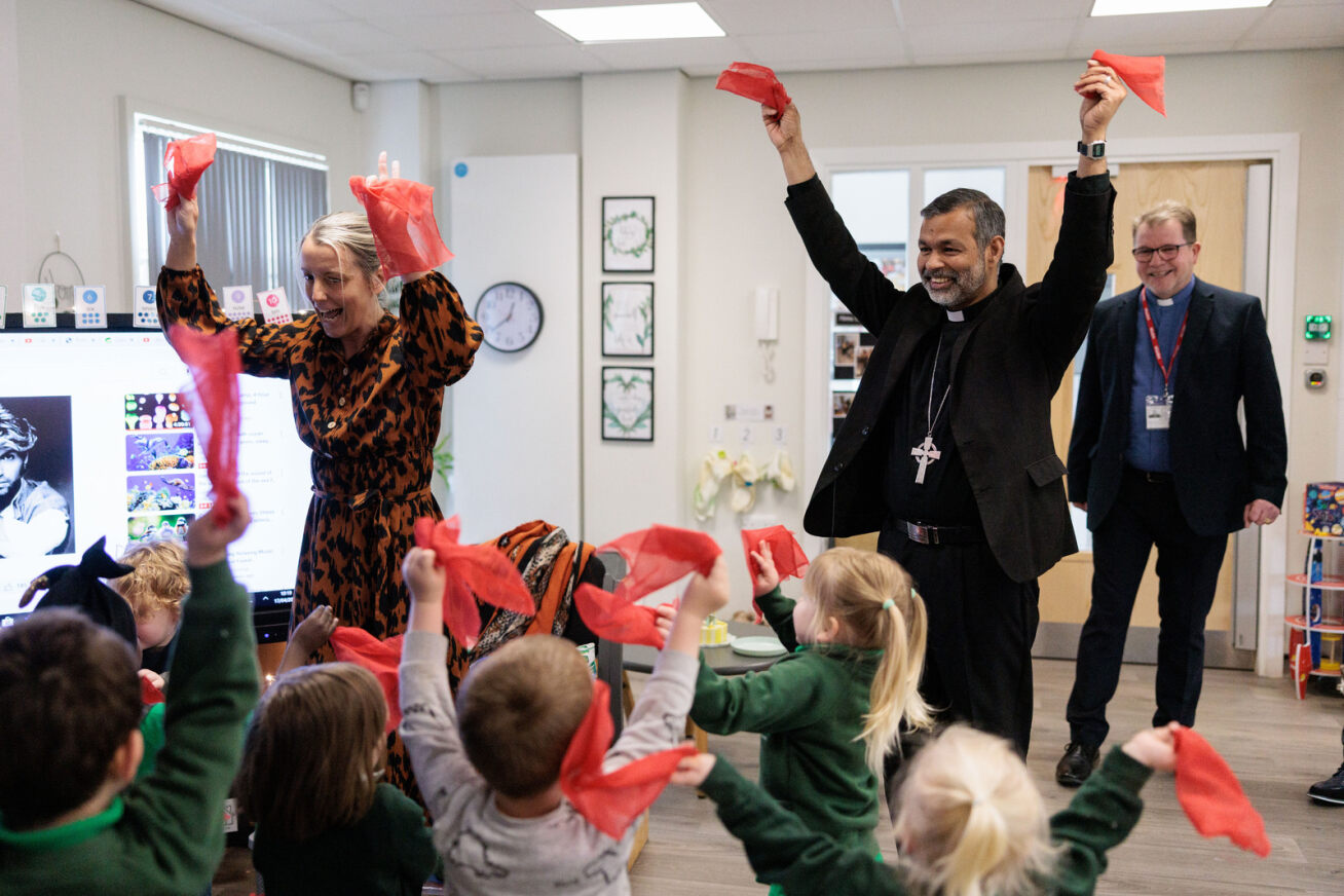 Bishop John in a primary school classroom with teacher and children all waving red flags. The bishop is joining in